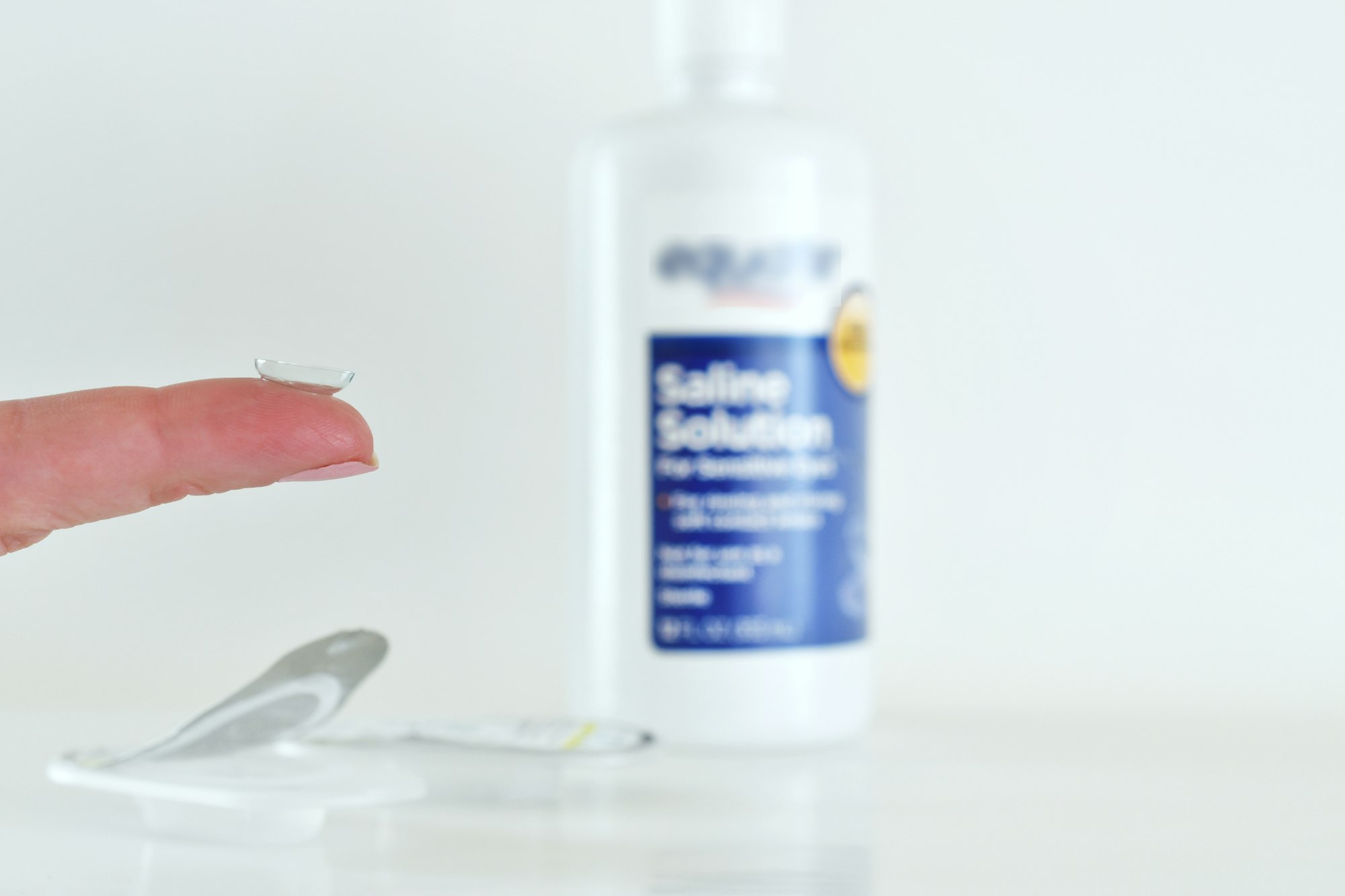 Female holding contact lens on her finger with disposable blister packs & saline solution bottle